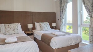 A hotel room with two beds, each with a headboard, bedside tables, lamps, and a view of the outdoors through a window with curtains.