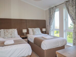 A hotel room with two beds, each with a headboard, bedside tables, lamps, and a view of the outdoors through a window with curtains.