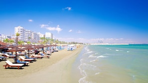 Une plage avec des transats et des parasols, un ciel dégagé et des bâtiments en arrière-plan.