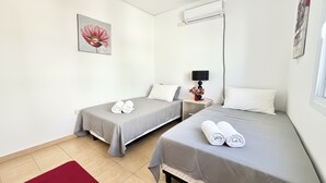 A hotel room with two single beds, a red rug, a wall-mounted air conditioner, and a painting of a flower. - 38 AN - Stay Malanda Sun Apartment (Ayia Napa)