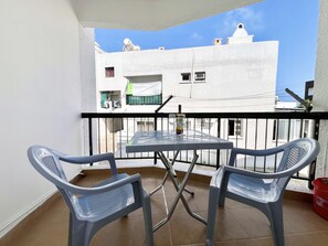 A balcony with a table and two chairs, a red bucket, and a white building in the background. - 38 AN - Stay Malanda Sun Apartment (Ayia Napa)