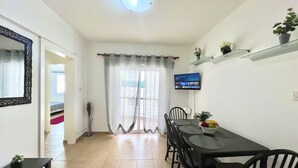 A dining area with a table and chairs, a television mounted on the wall, and a potted plant on a shelf. - 38 AN - Stay Malanda Sun Apartment (Ayia Napa)