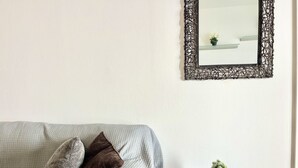 A living room with a grey sofa, two pillows, a small table with a plant, and a decorative mirror on the wall. - 38 AN - Stay Malanda Sun Apartment (Ayia Napa)