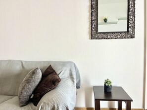 Ein Wohnzimmer mit grauem Sofa, zwei Kissen, einem kleinen Tisch mit Pflanze und einem dekorativen Spiegel an der Wand.
