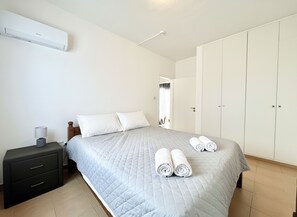 A bedroom with a bed, a nightstand, a wardrobe, and a window. - 38 AN - Stay Malanda Sun Apartment (Ayia Napa)