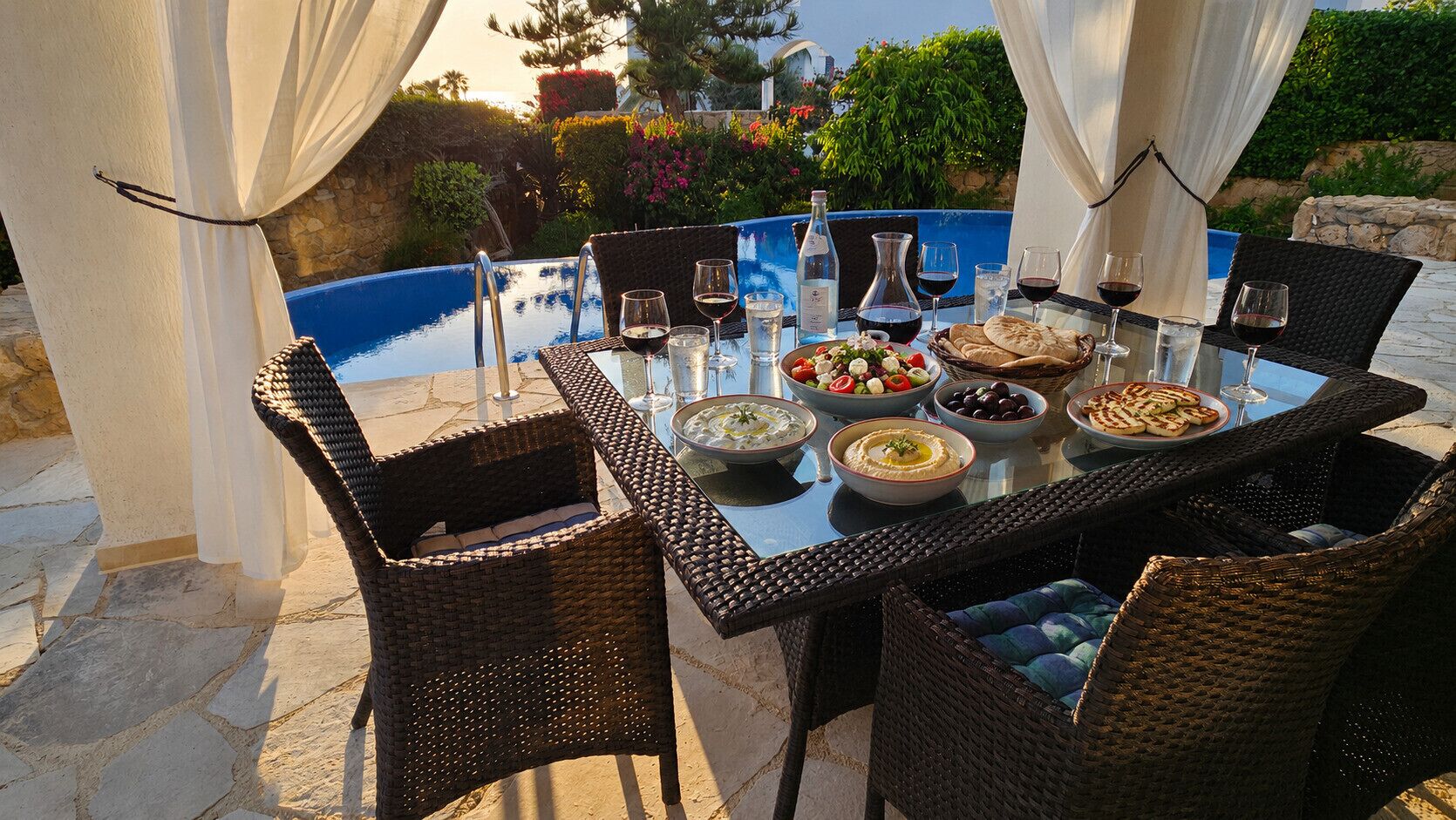 An outdoor dining area with wicker chairs, a glass table, and a pool in the background.