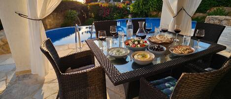 An outdoor dining area with wicker chairs, a glass table, and a pool in the background.