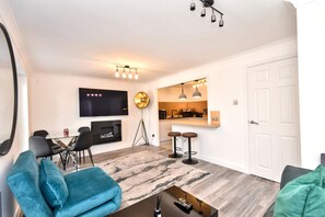 Ein modernes Wohnzimmer mit Flachbildfernseher, Kamin, Essbereich und einer Küche mit Pendelleuchten.