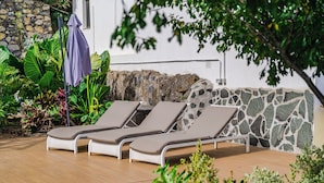 A sunlit outdoor seating area with three lounge chairs and a covered umbrella, surrounded by lush greenery and a stone wall. - Casa de Limon (La Vega)