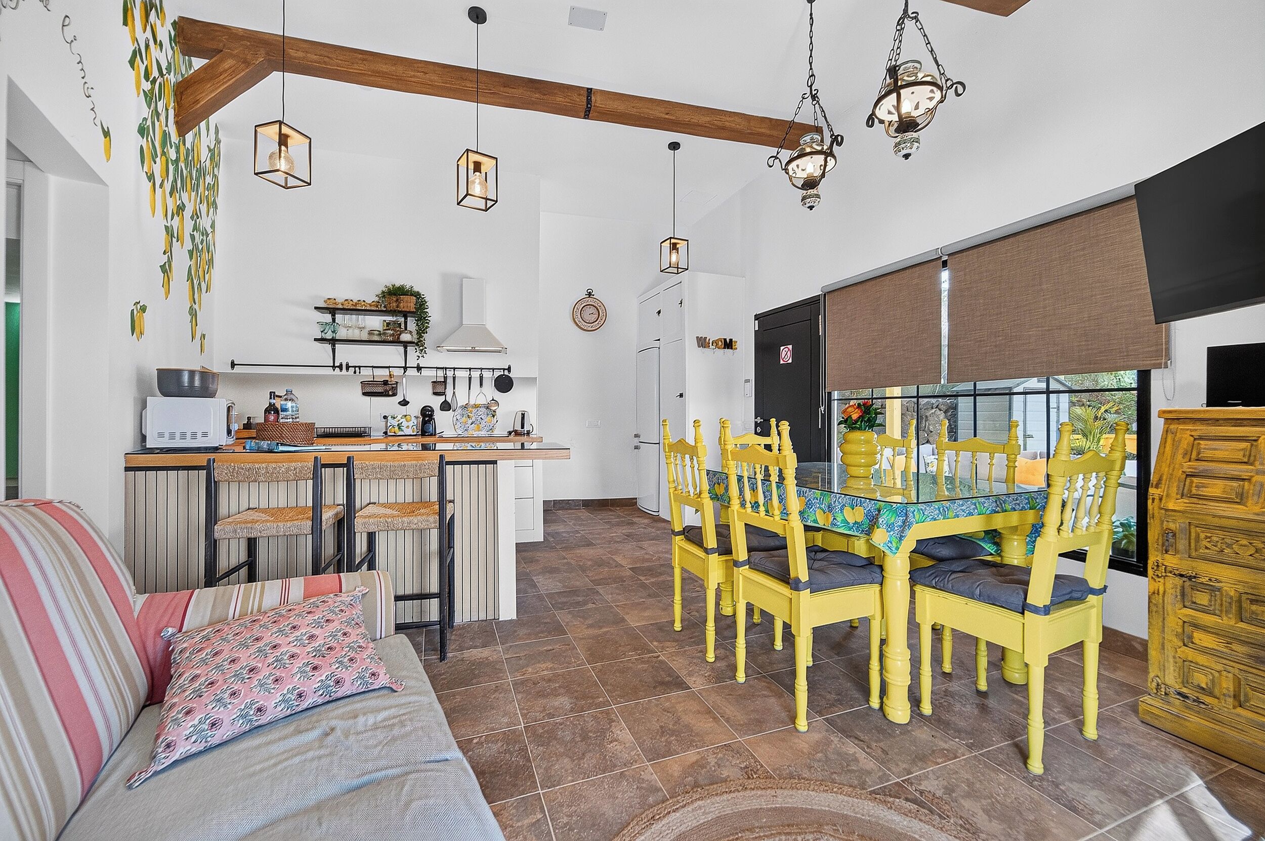 A modern kitchen with a dining area, featuring a central island, hanging lights, and a view through a large window.
