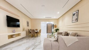 Una sala de estar moderna con televisor de pantalla plana, un sofá color beige, una zona de comedor con mesa y sillas, y un pasillo que lleva a una puerta.