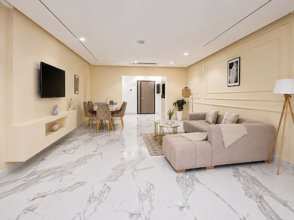 A modern living room with a sofa, a dining table with chairs, a flat-screen TV mounted on the wall, and a hallway leading to a door.