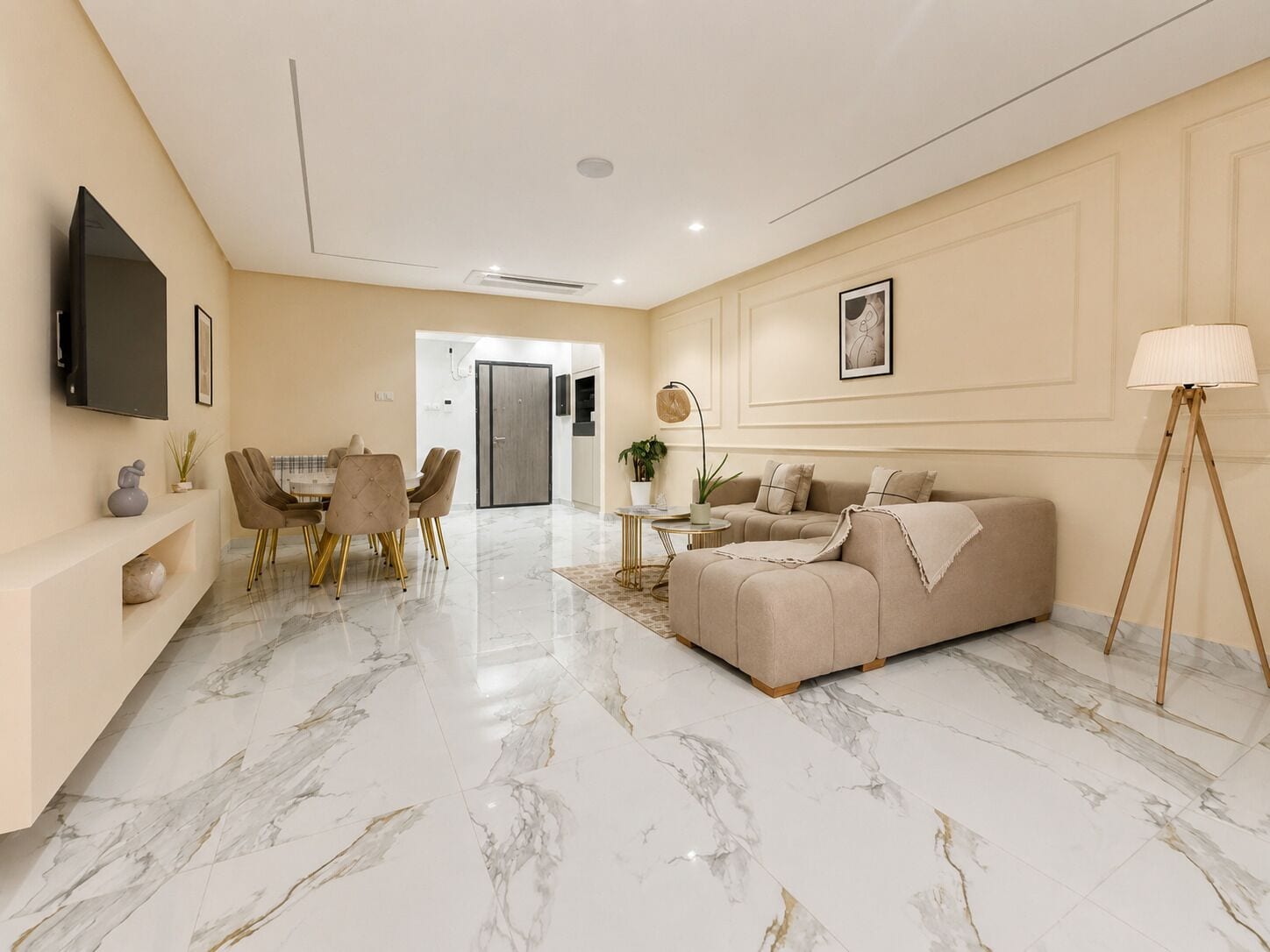 A modern living room with a marble floor, a beige sofa, a dining area with chairs, a TV mounted on the wall, and a lamp on a stand.