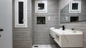 Un baño moderno con un tocador blanco, un grifo negro y suelo de mármol. Hay dos ventanas pequeñas con marcos blancos y un espejo sobre el tocador.