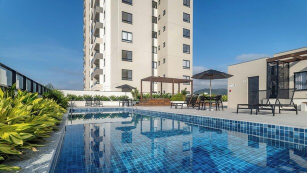 Ein Schwimmbecken mit Fliesenrand, umgeben von Liegestühlen und Sonnenschirmen, mit einem hohen Gebäude im Hintergrund.