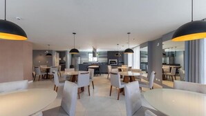 A modern dining area with multiple tables and chairs, a kitchen in the background, and pendant lighting. - Studio in a Cond. With a gym in Downtown Muv0811 (Curitiba)