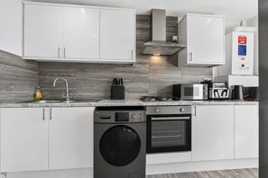 Una cuina moderna amb armaris blancs, una campana d'aspiració d'acer inoxidable, un microones integrat, una cafetera i una rentadora.