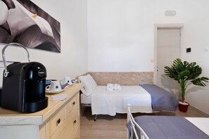 A hotel room with a bed, a desk with a coffee maker, a plant, and a painting on the wall.