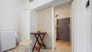 Une pièce de taille modeste, avec une chaise blanche, une petite table sur laquelle se trouve un bol, une armoire blanche et un espace douche avec un mur recouvert de carreaux.