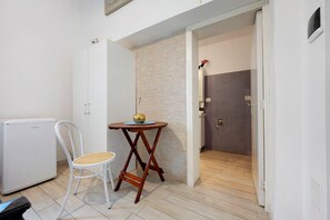 Une pièce de taille modeste, avec une chaise blanche, une petite table sur laquelle se trouve un bol, une armoire blanche et un espace douche avec un mur recouvert de carreaux.