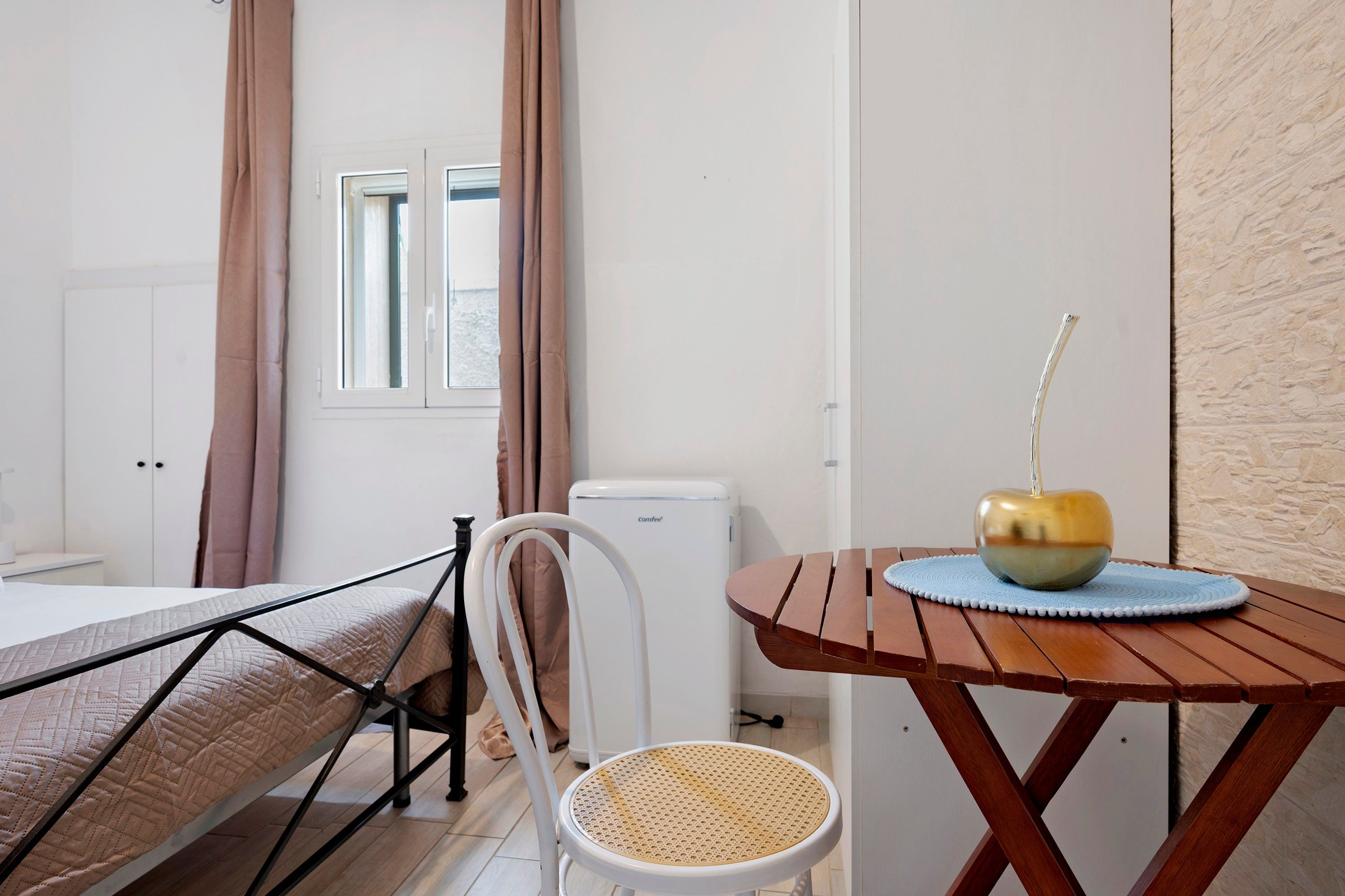 Een kamer met een bed, een stoel, een tafel met een decoratieve schaal en een raam met gordijnen.