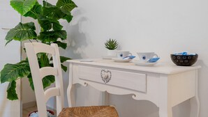 Un bureau blanc avec un tiroir, une petite plante en pot, une tasse blanche ornée d’un logo bleu, une soucoupe blanche, un panier noir et une chaise en osier.
