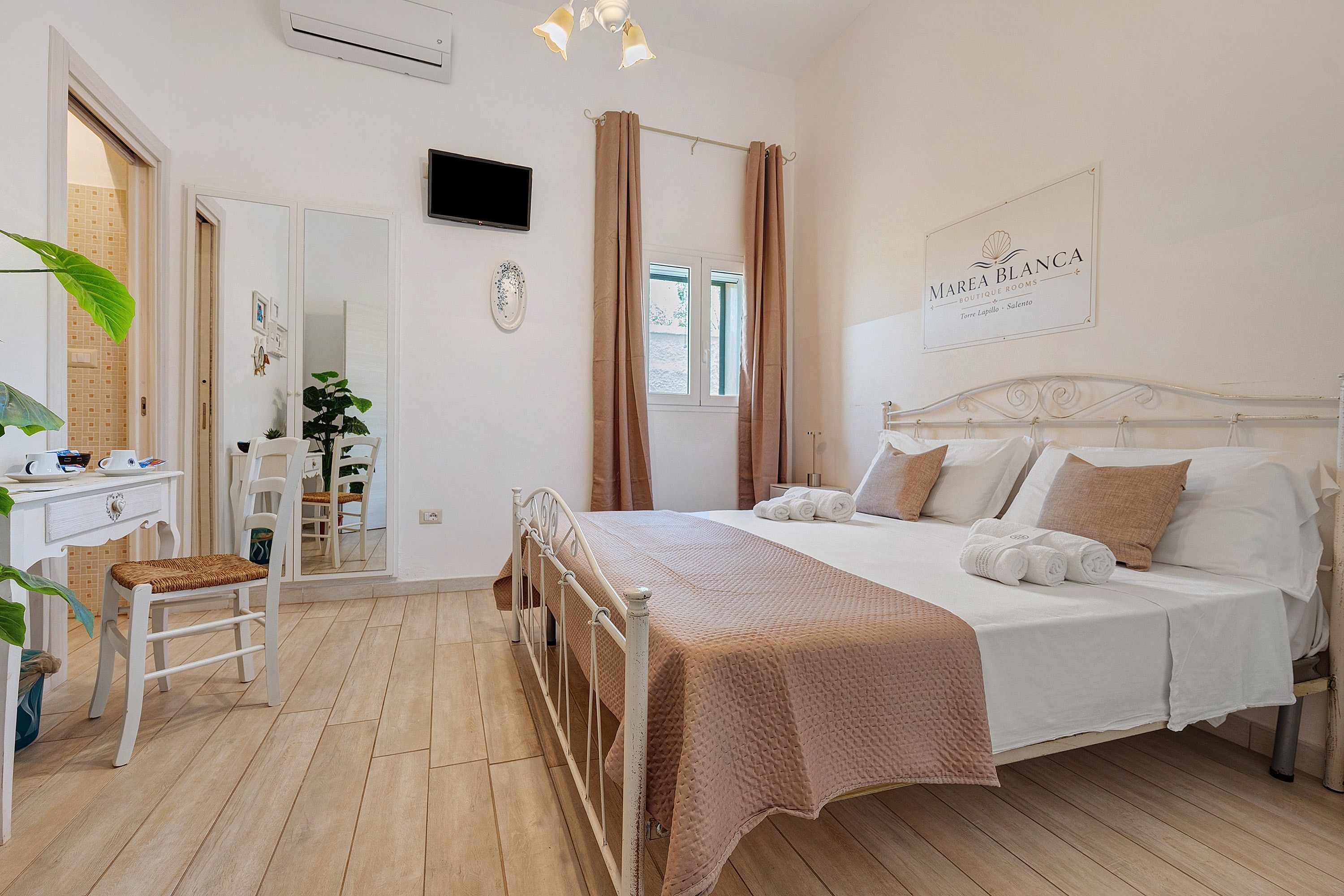 Une chambre d’hôtel avec un lit, une table de chevet et un bureau avec une chaise. Il y a une fenêtre avec des rideaux, un téléviseur fixé au mur et une enseigne indiquant le nom de l’hôtel.