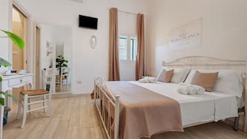 Ein Hotelzimmer mit Bett, Nachttisch und Schreibtisch mit Stuhl. Es gibt ein Fenster mit Vorhängen, einen an der Wand montierten Fernseher und ein Schild mit dem Namen des Hotels.