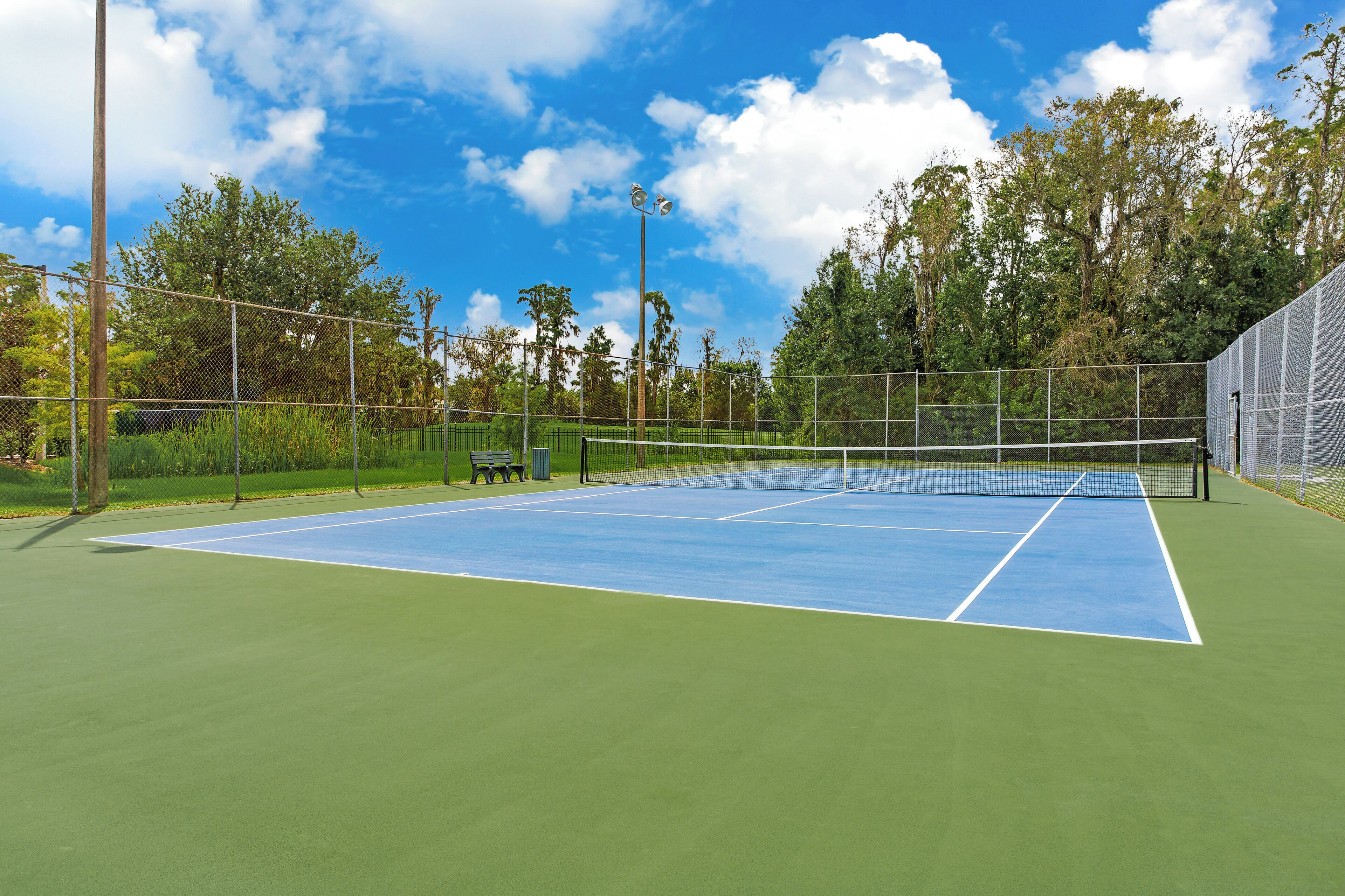 An outdoor tennis court with a net, surrounded by a chain-link fence, and trees in the background.
