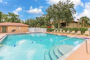 A swimming pool with a white fence, lounge chairs, and a building with a red roof. - Your Florida Launch Pad | Pool Days & Sunshine Stays (Kissimmee)