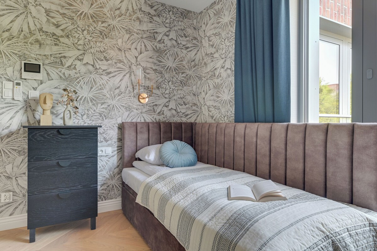 Un dormitorio con cama, cómoda, luz empotrada en la pared y ventana con cortinas.