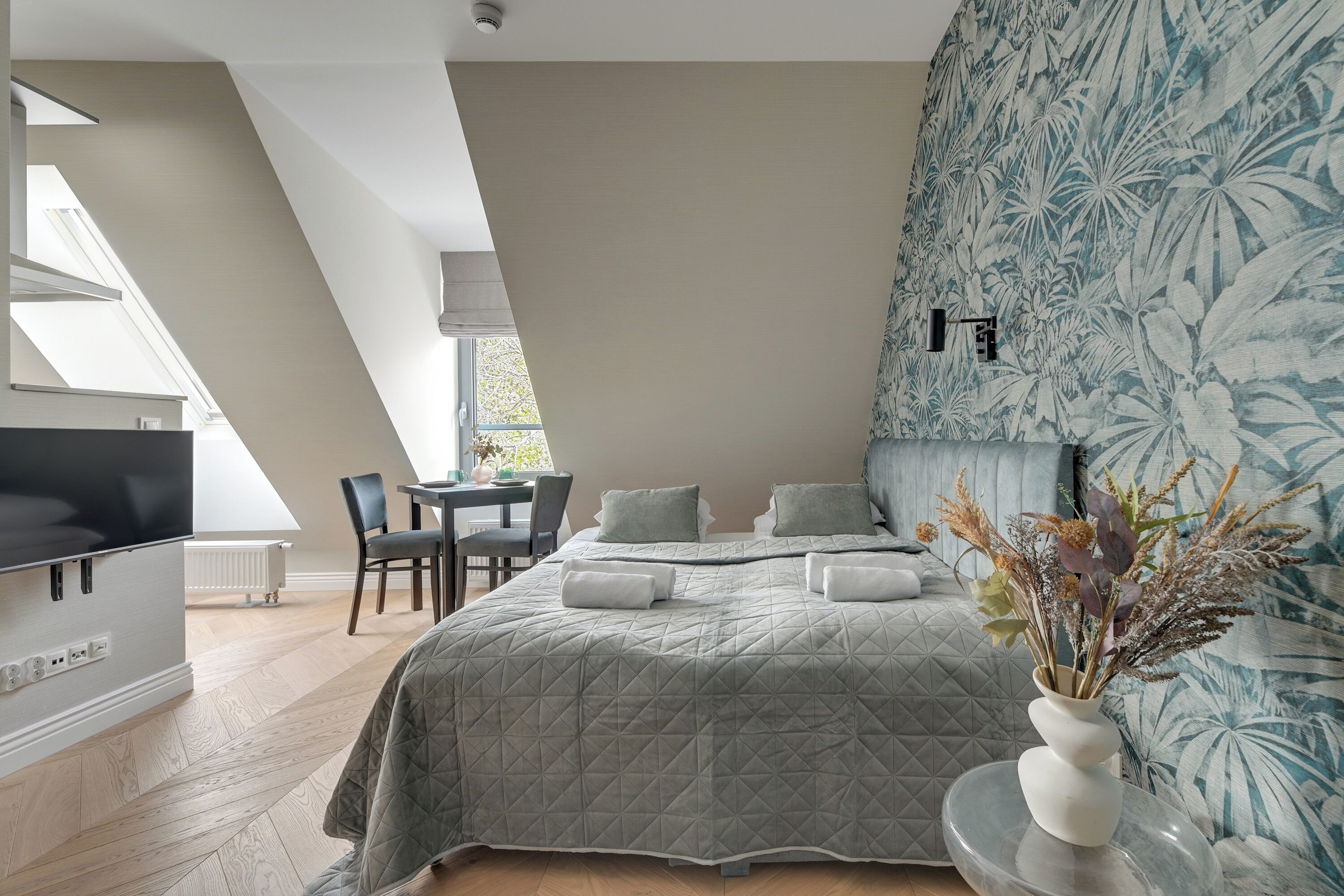 A modern bedroom with a large bed, a TV, a dining area, and a window with a view.