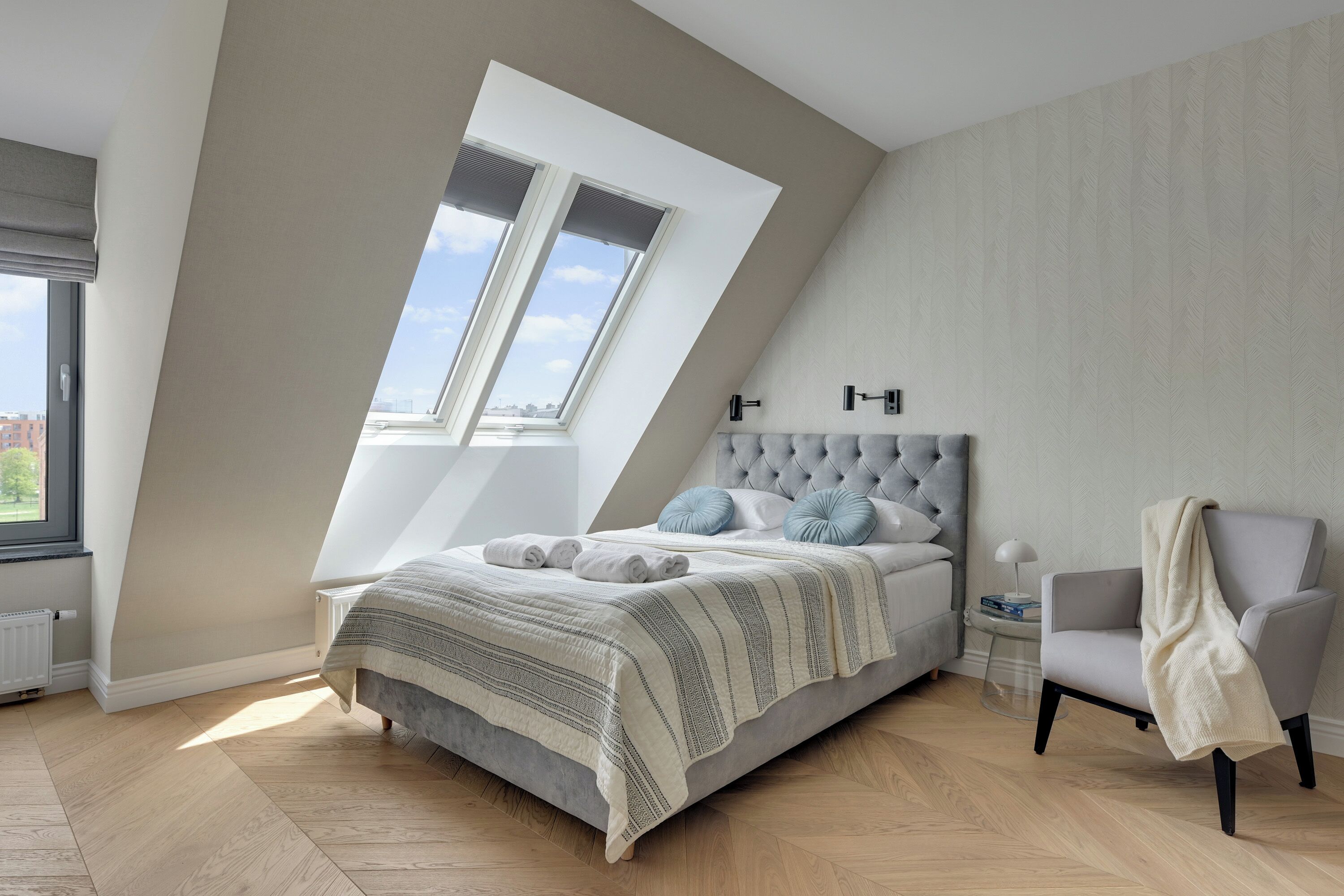 Un dormitorio con techo inclinado, una cama con cabecera capitonada, una silla y una ventana con persianas.
