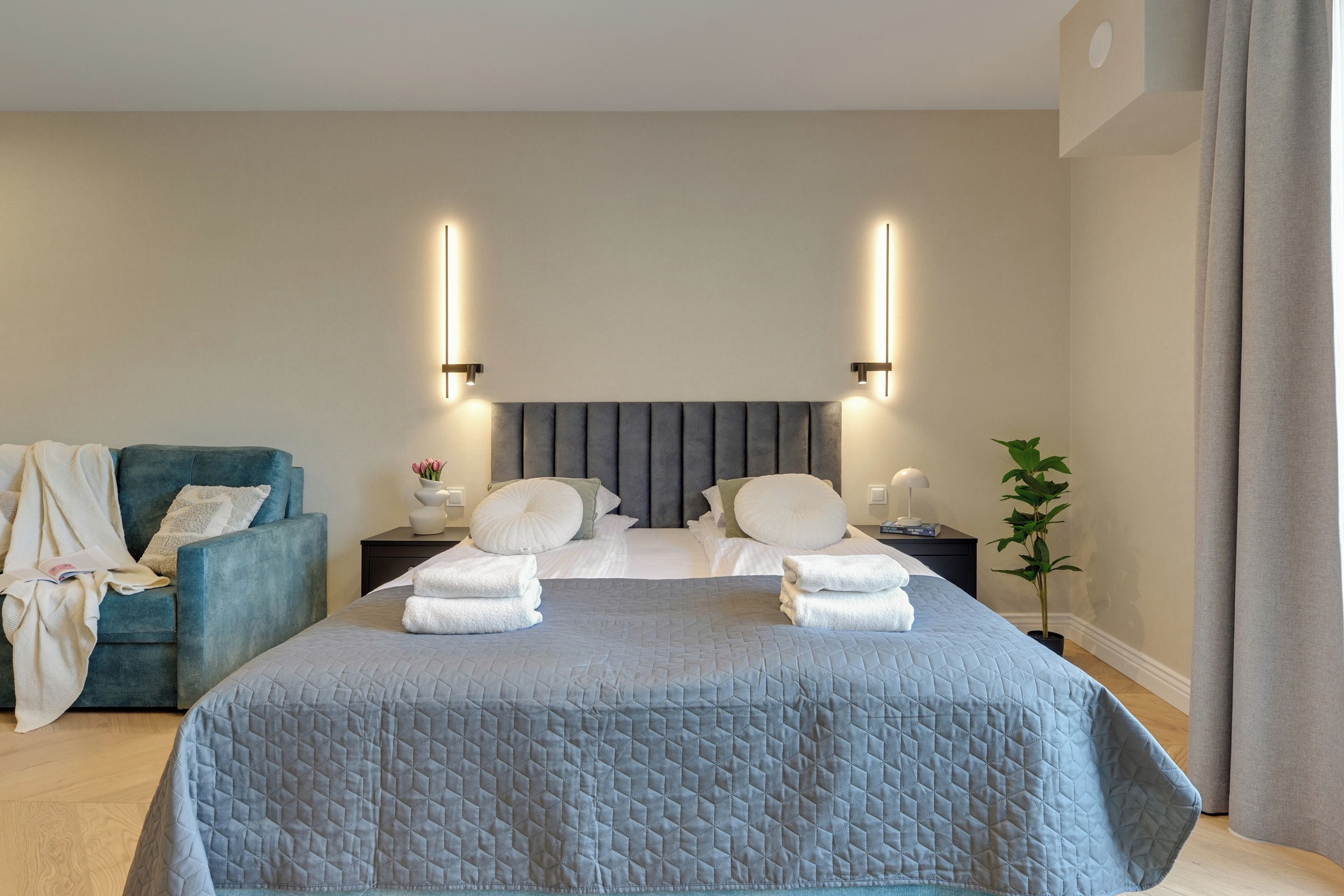 Une chambre d’hôtel avec un grand lit, un fauteuil bleu, une table de chevet avec une lampe et une plante en pot.