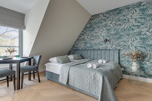 Un dormitorio con techo inclinado, una cama con colcha acolchada, un escritorio con silla y una ventana con vistas a árboles.