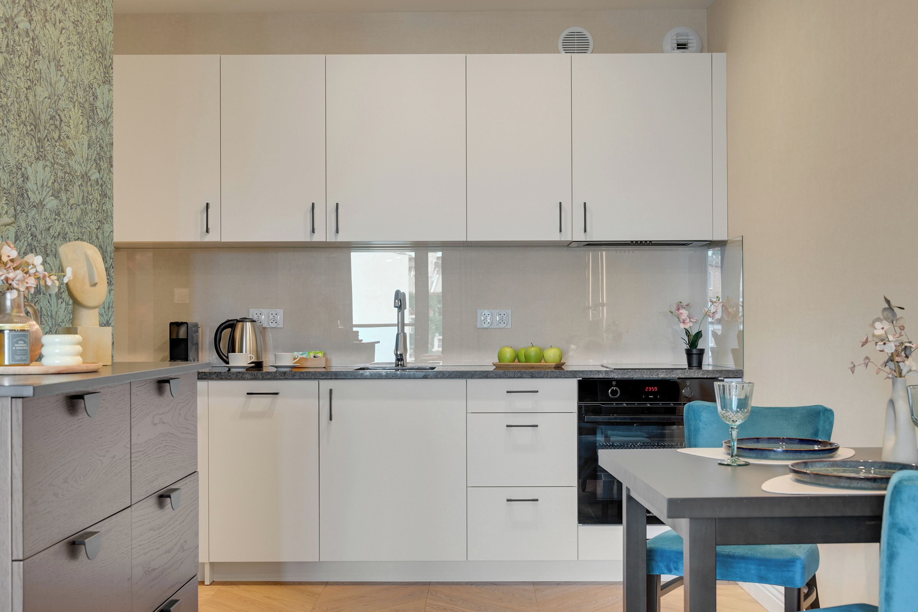 A modern kitchen with white cabinets, a built-in oven, and a dining area with a table and chairs.
