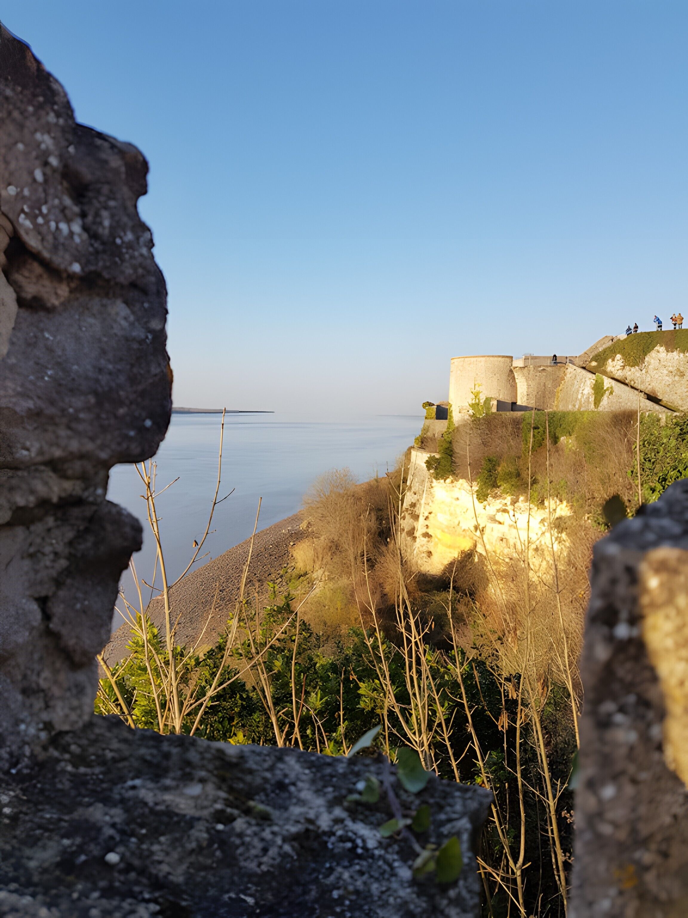 A coastal view with a stone structure overlooking the sea, vegetation in the foreground, and a clear sky.