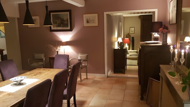Comedor con mesa de madera, sillas e iluminación colgante. Se observa una puerta que da a otra habitación con un sofá y una lámpara.