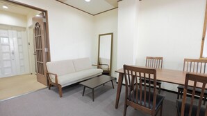 Een kamer met een bank, een tafel en stoelen. Er is een deur met een raam en een spiegel aan de muur.