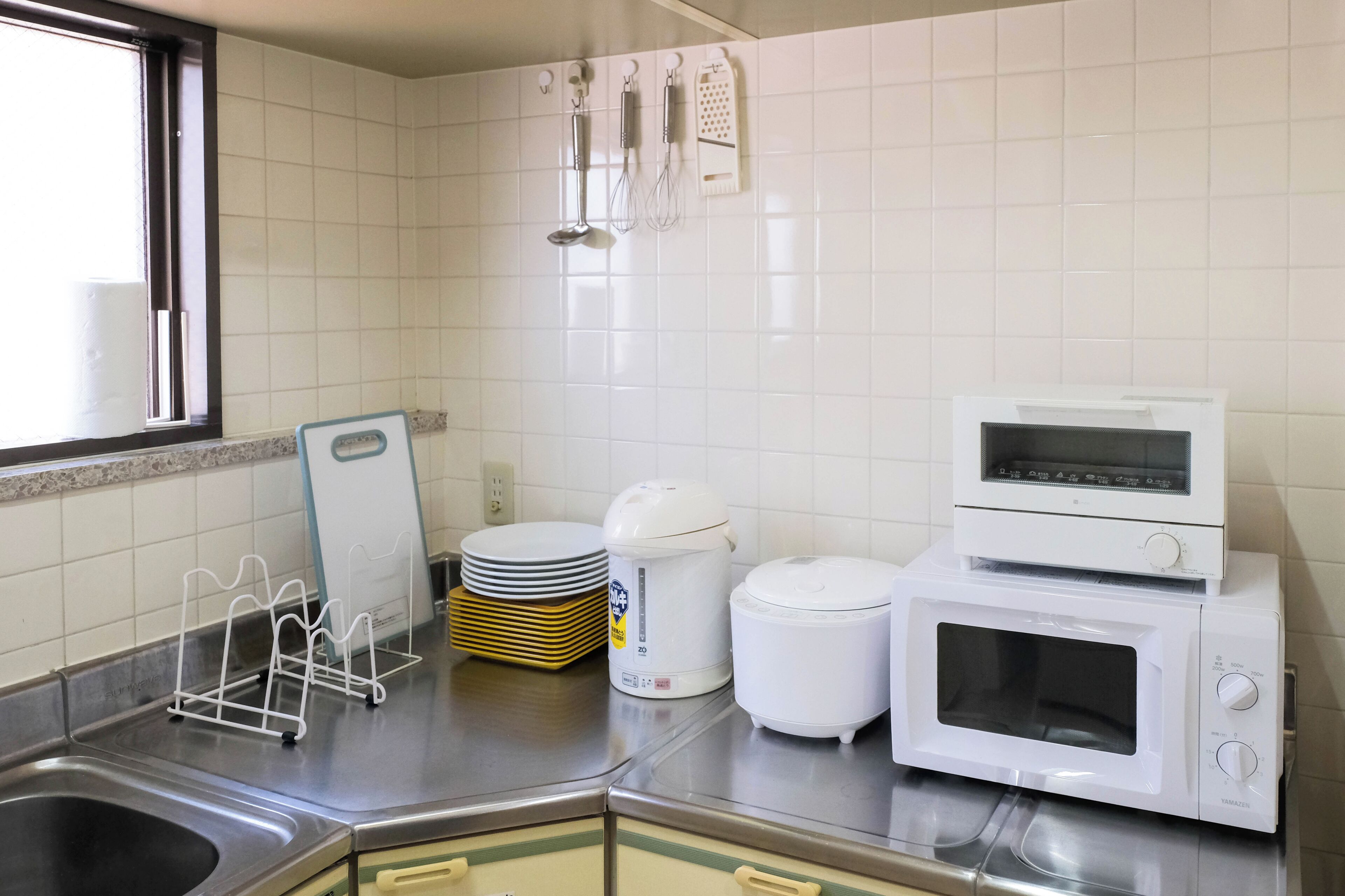 A kitchen with a stainless steel sink, a microwave, a rice cooker, and a stack of plates.