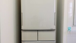 Een witte koelkast met een vriesvak bovenop en een tweede vriesvak onderin, geplaatst in een hoek van een kamer met witte muren en een betegelde vloer.