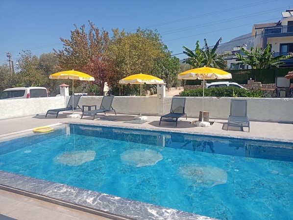 A swimming pool with lounge chairs and umbrellas, surrounded by a low wall and vegetation.