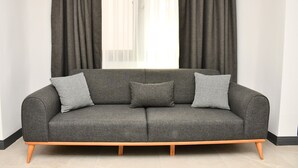A grey sofa with wooden legs and houndstooth pillows, positioned in front of a window with sheer curtains. - Sönmez Güler Pansiyon Ölüdeniz (Muğla)