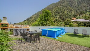 Een terras met een tafel en stoelen, een barbecue, een blauw zwembad en uitzicht op een beboste heuvel.