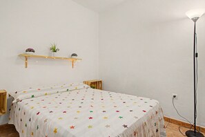 A room with a bed, a bedside table, a wall-mounted shelf with plants, and a floor lamp. - Casa Levante Conil – Cozy Apartment in the Heart of Conil de la Frontera (Conil de la Frontera)