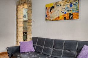 Ein Zimmer mit einer schwarzen Ledersofa, zwei violetten Kissen, einer Ziegelwand und einem farbenfrohen Gemälde an der Wand.