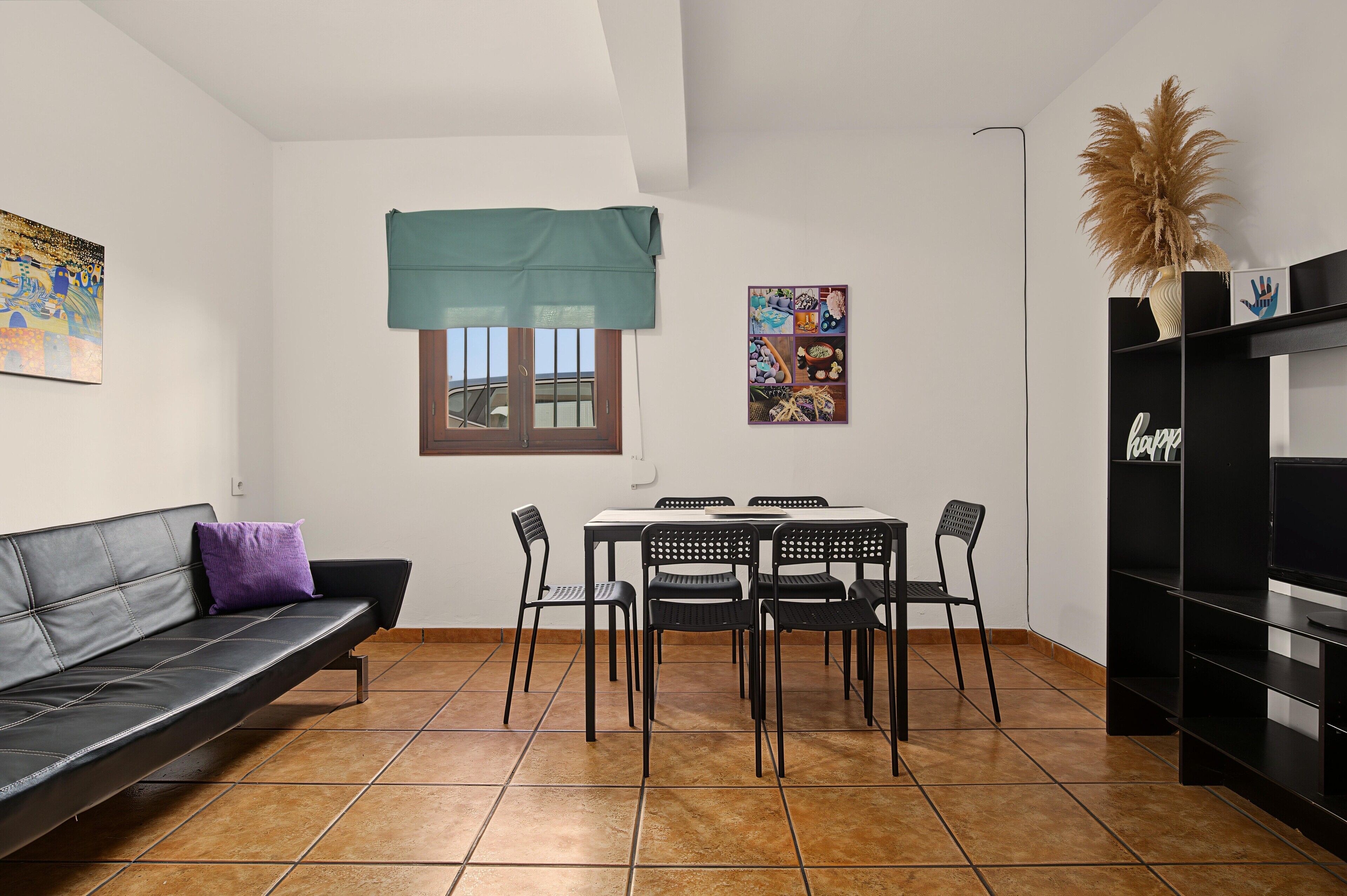 A compact living space with a sofa, dining table, chairs, a small window with a curtain, and a wall-mounted shelf with decorative items.