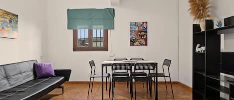 A compact living space with a sofa, dining table, chairs, a small window with a curtain, and a wall-mounted shelf with decorative items.