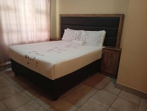 Un dormitorio con cama, cabecera, mesita de noche y ventana con cortinas.