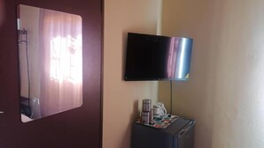 Una habitación con un televisor montado en la pared, un refrigerador pequeño con una bandeja de objetos encima y una puerta con un espejo que refleja parte de la habitación.